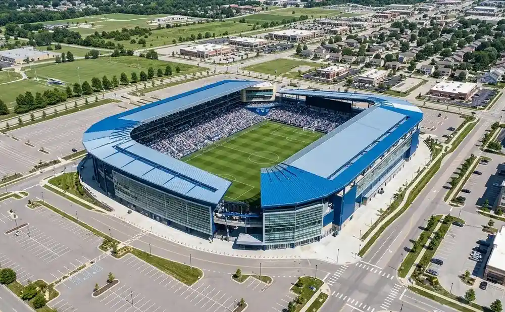 Aerial view of a soccer stadium with blue roofing, surrounded by parking lots and residential and commercial buildings.