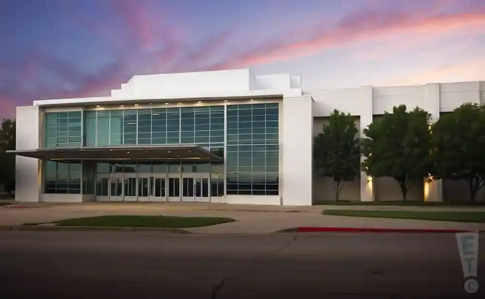 Modern white building with large glass windows and a canopy over the entrance, under a colorful sunset sky.