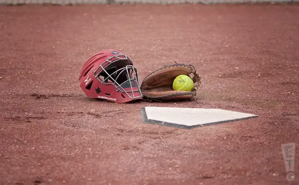 A catcher's mask and mitt with a softball sit on a baseball field next to home plate.