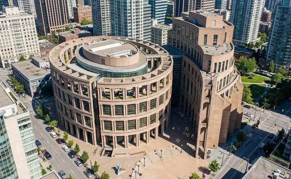Aerial view of a circular, Brutalist library with beige columns, surrounded by modern skyscrapers.