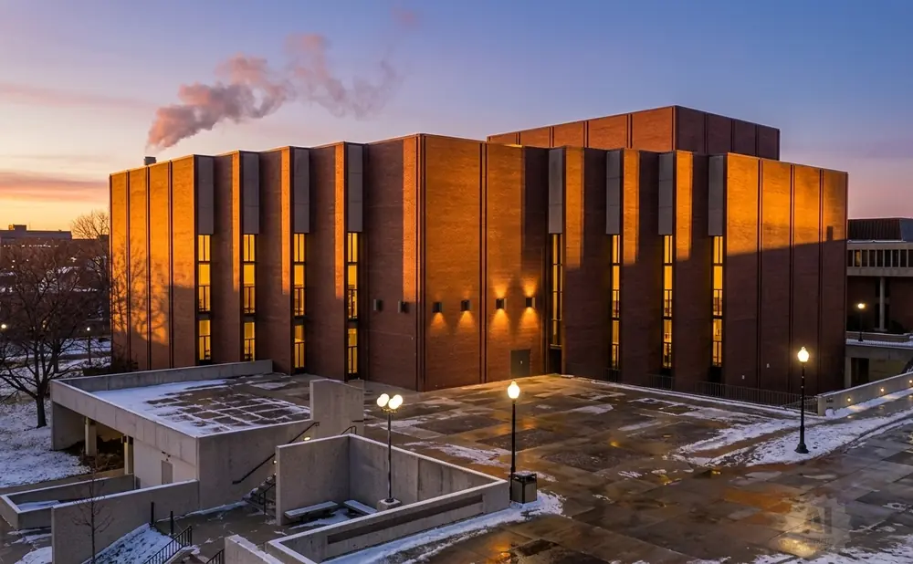 Sunset illuminates a modern brick building with vertical windows, steam rises from a chimney.