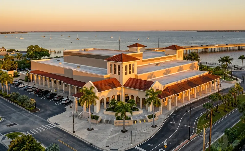 An aerial view of a large, tan building with a red tile roof, resembling a theater or convention center, with palm trees and a parking lot.