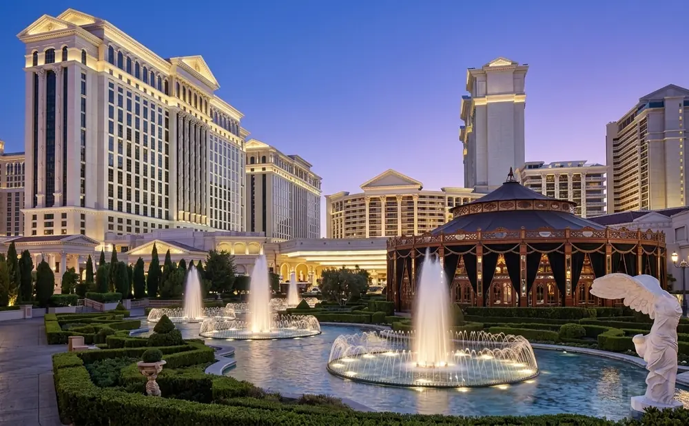 Fountains and lush gardens in front of a grand Roman-style hotel and casino at dusk.