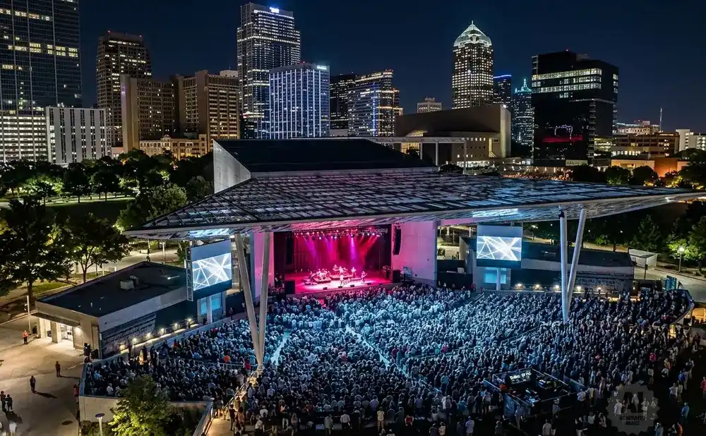 An outdoor concert at night with a band on stage, a large crowd, and a city skyline in the background.