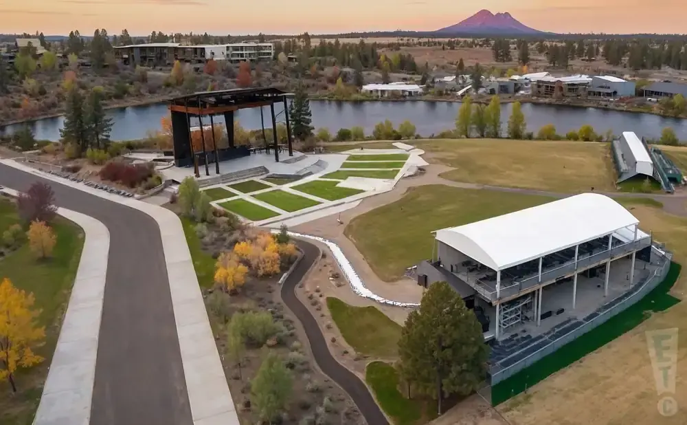 Aerial view of an outdoor amphitheater with tiered seating, a stage, and a large tent venue near a lake.