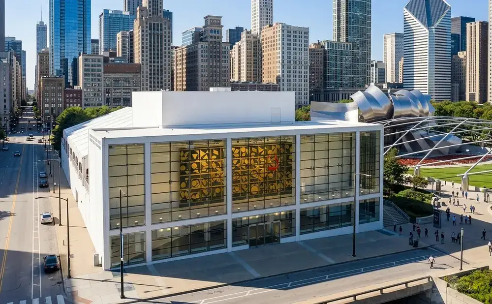 Modern white building with large glass windows displaying artwork, set against a cityscape and park.