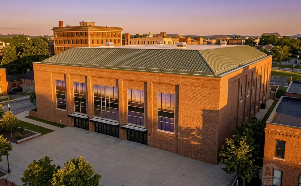A large brick building with a green roof and tall windows reflecting the sunset, viewed from above.