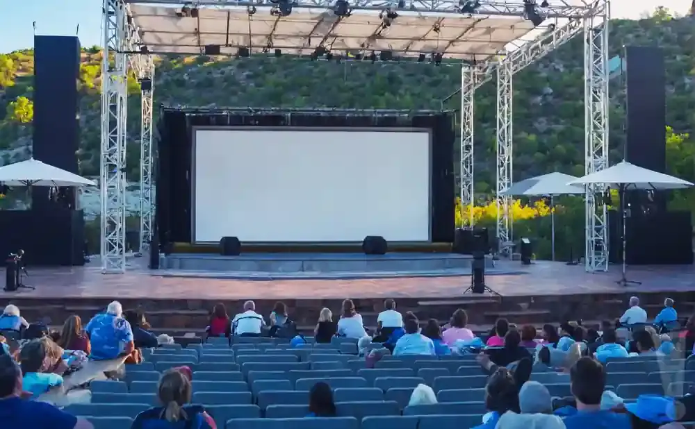 Outdoor movie screen with tiered seating for an audience in a natural setting.