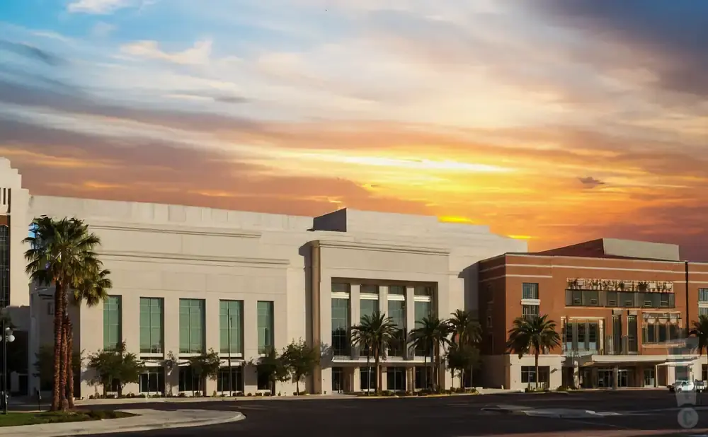 A modern building with a tan facade and large windows, surrounded by palm trees, under a dramatic sunset sky.