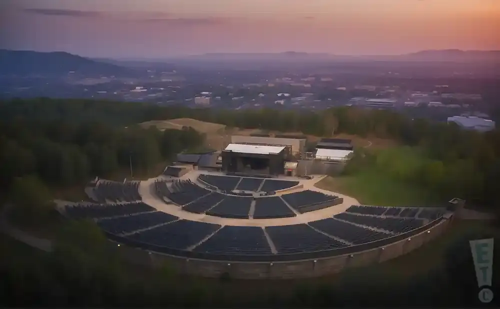 Aerial view of an outdoor amphitheater with tiered seating, a stage, and a city skyline in the background at dusk.