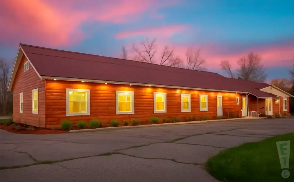 Long, red-brown wooden building with many lit windows at dusk, under a pink and blue sky.