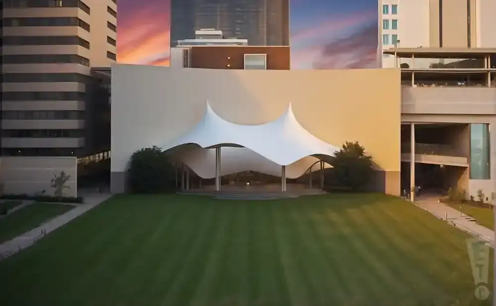 An outdoor amphitheater with a white tent roof and a grassy lawn in front, framed by modern buildings.