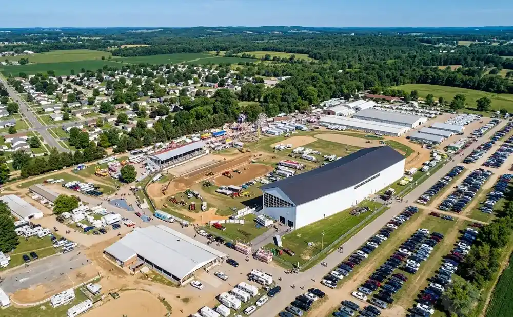 Aerial view of a fairground with buildings, parking lots, RVs, and a Ferris wheel under a blue sky.