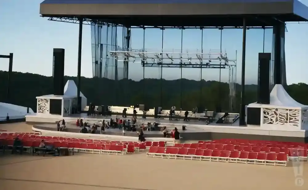 An outdoor amphitheater stage with empty red seats in the foreground and trees in the background.