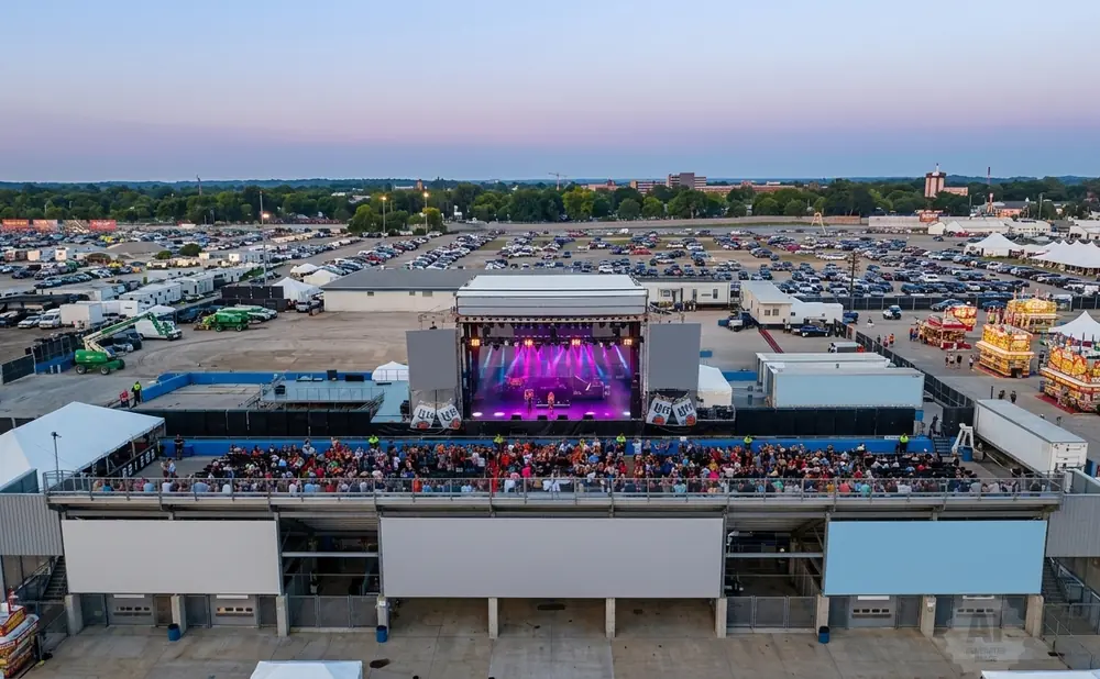 Aerial view of an outdoor concert stage with a band performing, facing a seated audience, with a large parking lot and fairgrounds in the background.