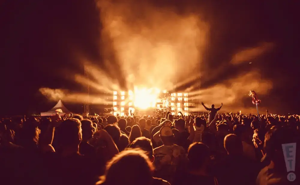 A crowd at a nighttime concert with bright stage lights and smoke.