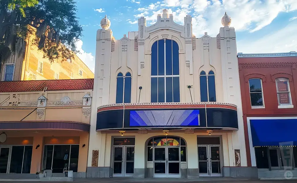 Art deco theater with a large marquee screen and multiple arched windows, under a cloudy blue sky.