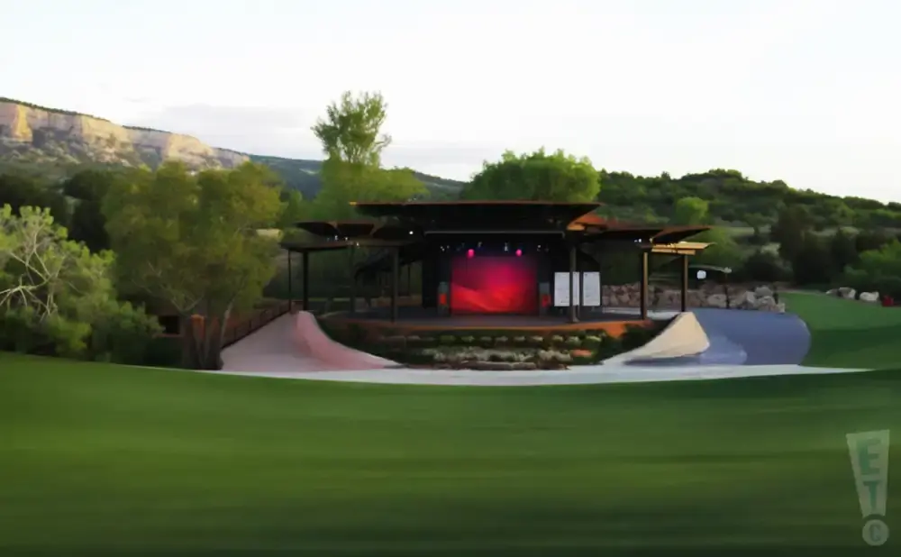 Outdoor amphitheater stage with a red backdrop, set against a landscape of green hills and a rocky cliff under a cloudy sky.
