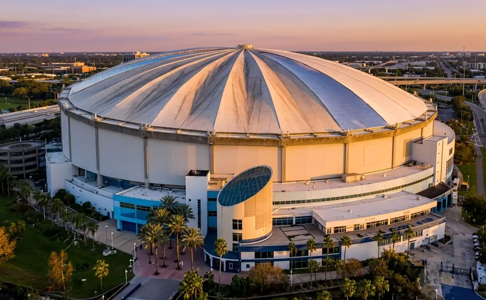 Aerial view of Tropicana Field, a domed stadium, at sunset with surrounding highways and palm trees.