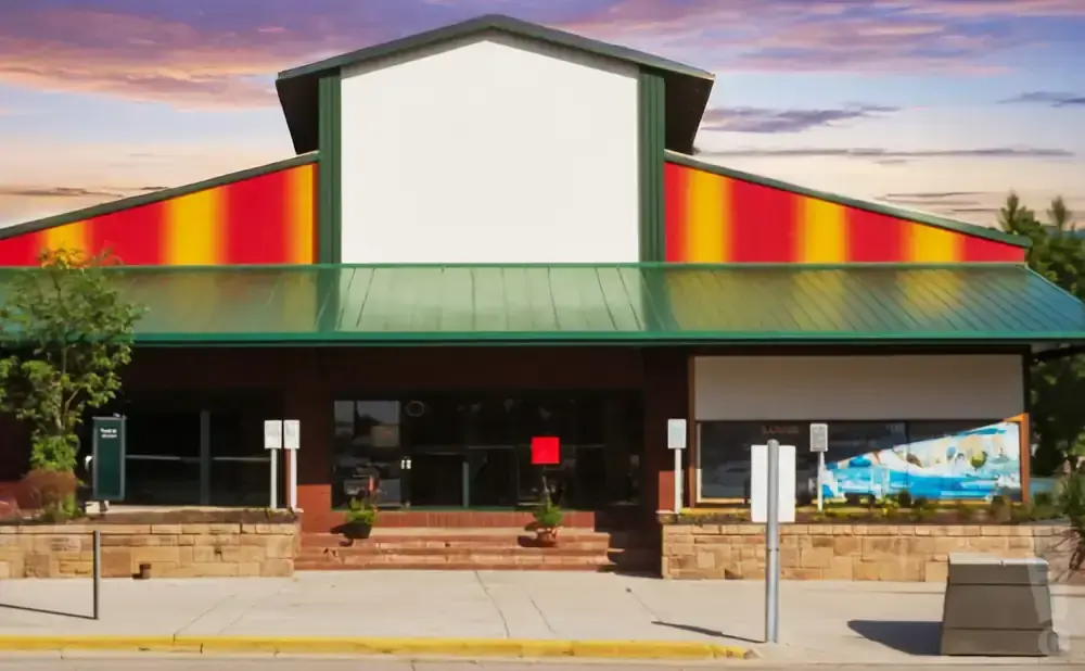 Building with a large blank white sign and red and yellow striped roof, with trees and a sidewalk in front.