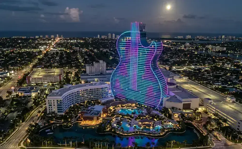 Aerial view of a guitar-shaped hotel lit with neon lights at night, overlooking a city and the ocean.