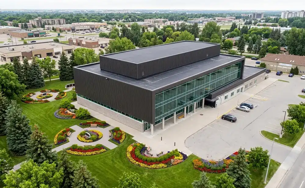 Aerial view of a modern, dark-paneled building with large glass windows, surrounded by lush green lawns and colorful flower beds.