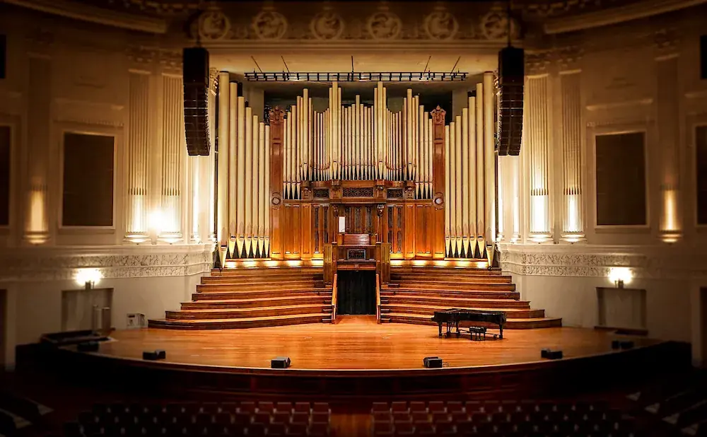A grand pipe organ dominates the stage of a concert hall, with a grand piano to the right.