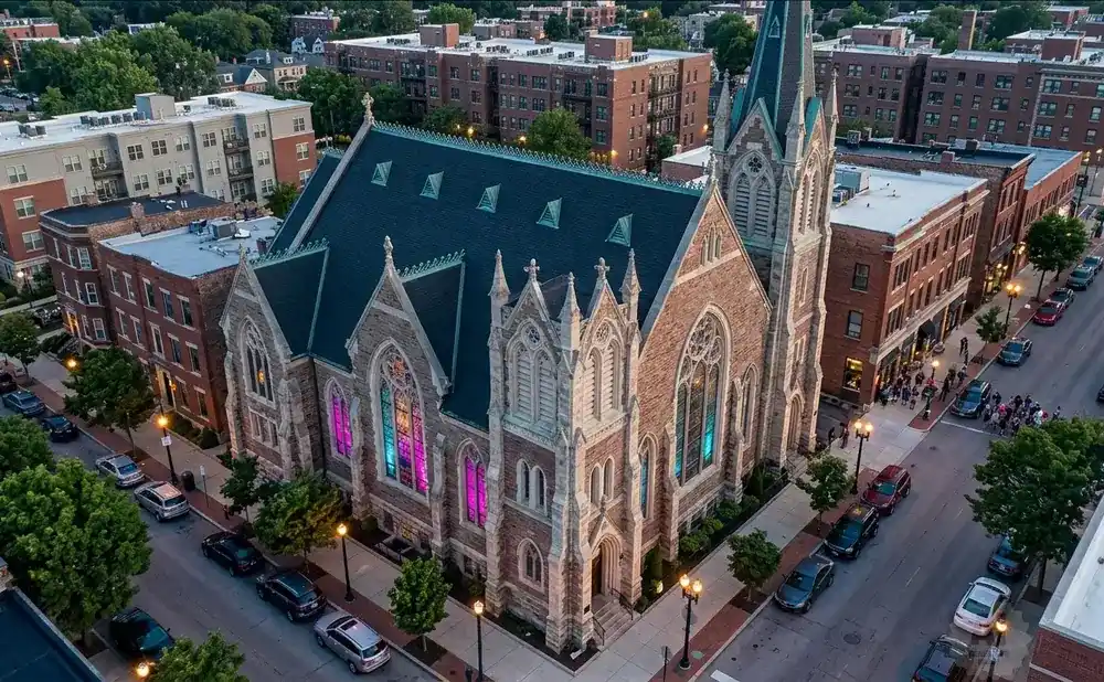 Aerial view of a stone church with stained glass windows illuminated in pink and blue, surrounded by brick buildings and trees.