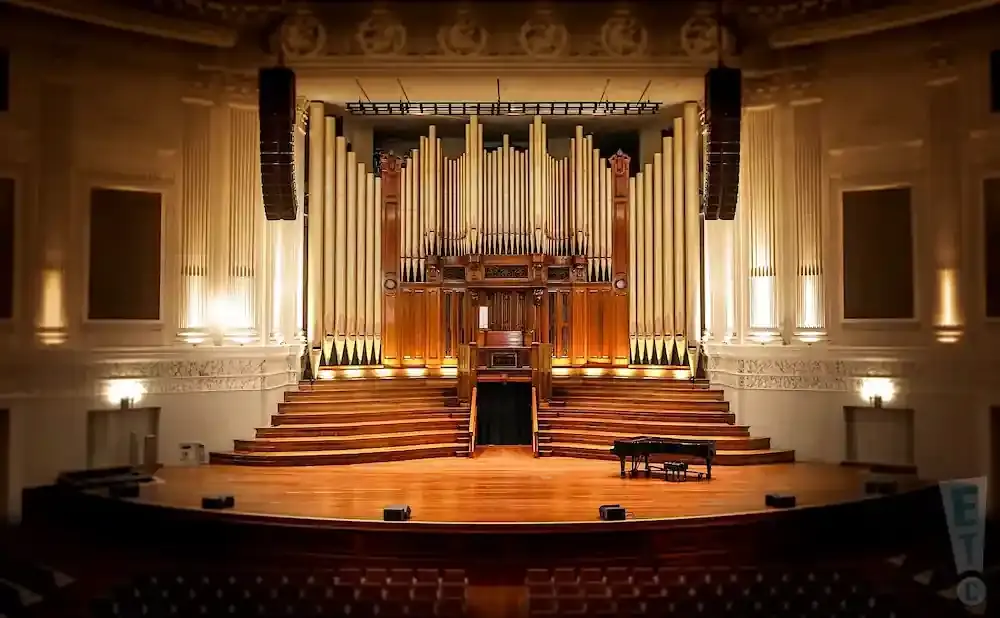 An empty concert hall stage with a grand piano, organ, and tiered seating.