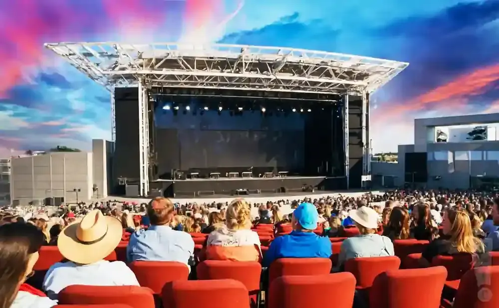 Outdoor concert stage with a large audience seated in red chairs. The sky is a vibrant mix of pink and blue.