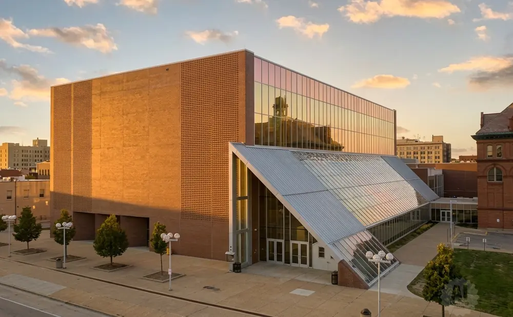 Modern brick and glass building with a dramatic slanted glass entryway, reflecting a cloudy sunset.