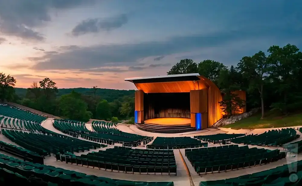 An outdoor amphitheater with green seats and a wooden stage is lit by the sunset.