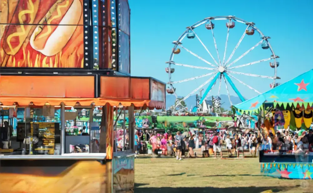 Carnival scene with a Ferris wheel, food stalls, and a crowd of people enjoying the sunny day.
