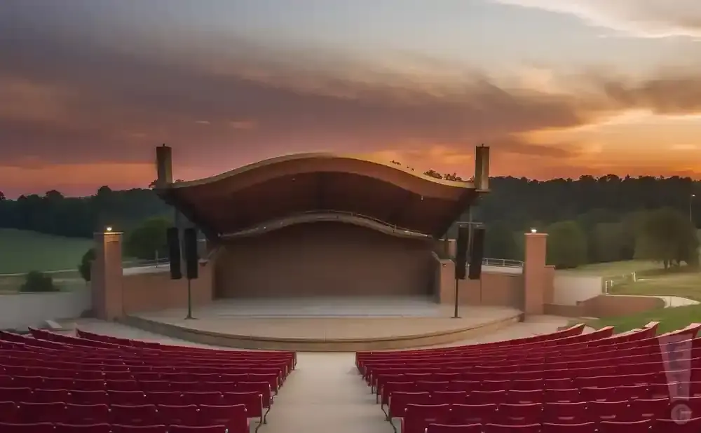 Outdoor amphitheater with red seats facing a stage under a colorful sunset sky.