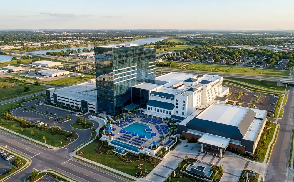 Aerial view of a modern resort with a tall glass building, outdoor pool, and surrounding cityscape.