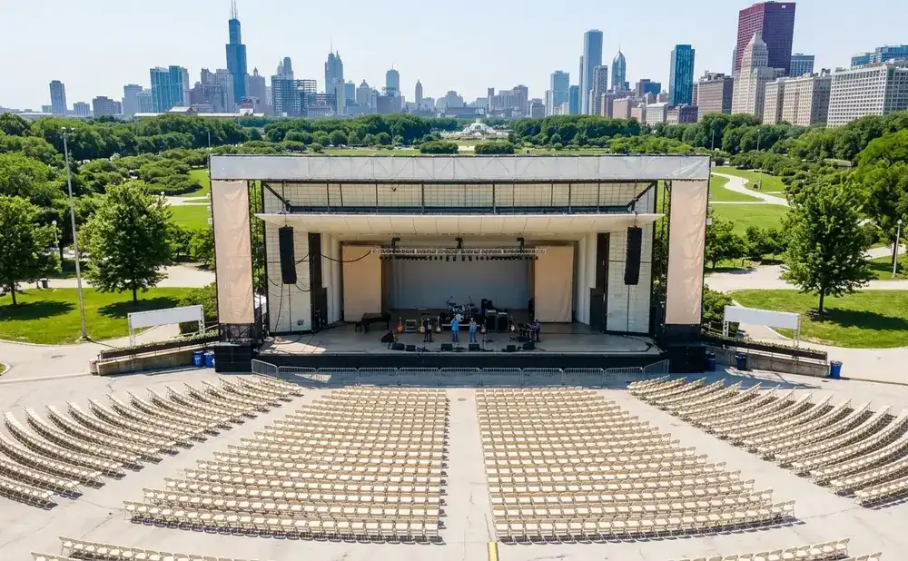 An empty outdoor concert venue in a park, with a stage set up and rows of beige chairs facing it.