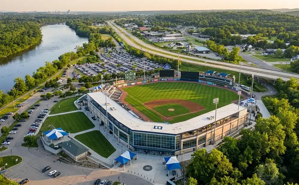 Aerial view of a baseball stadium surrounded by trees and a river, with a highway in the background.