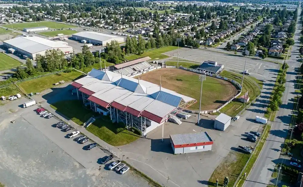 Aerial view of an arena with stadium seating and a dirt track, surrounded by sports fields, houses, and parking lots.