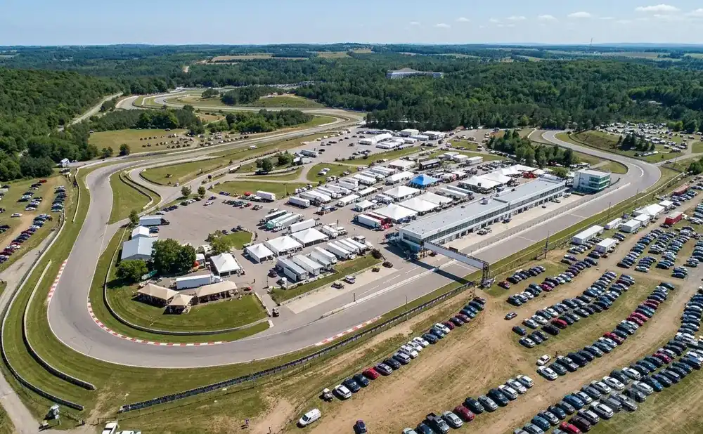 Aerial view of a race track with many cars parked in the paddock area, surrounded by green hills and trees.