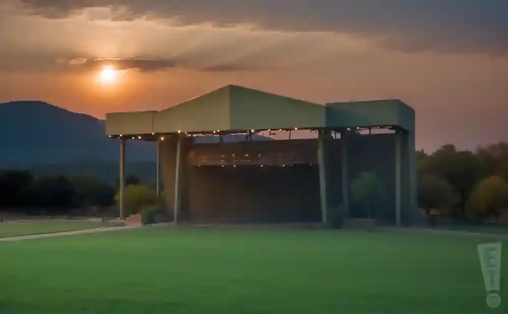 Outdoor amphitheater stage with a grassy lawn in the foreground, mountains and sunset in the background.