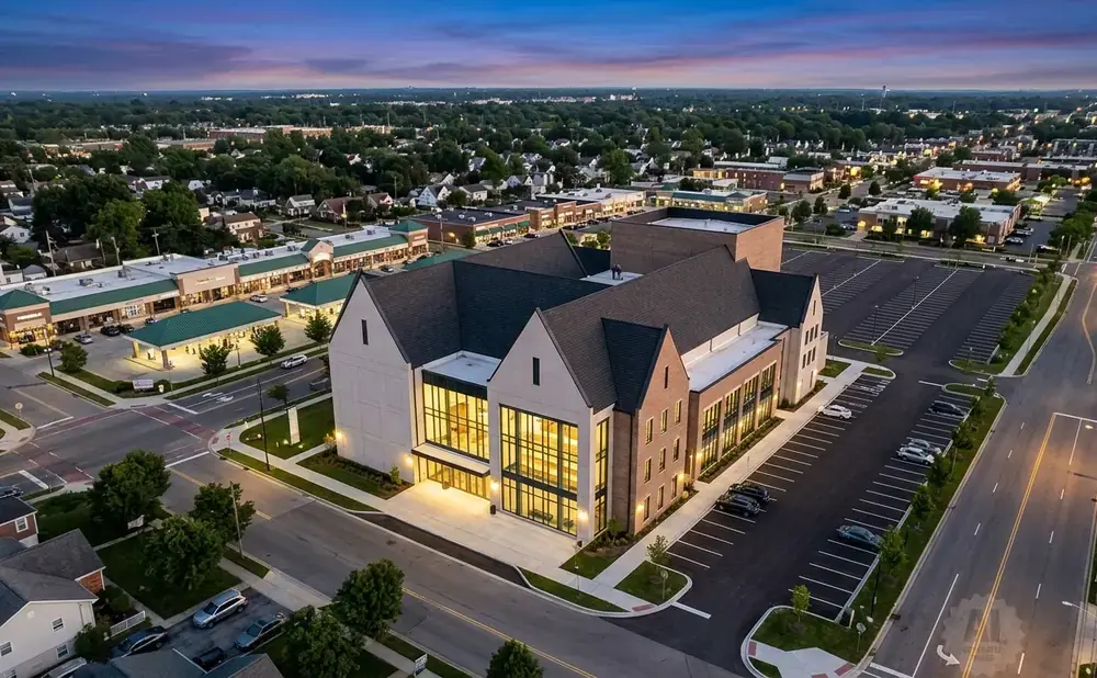 Aerial view of a modern church building with large windows and brick facade, surrounded by a suburban landscape and parking lot at twilight.