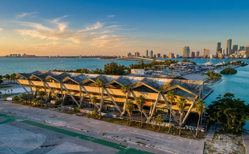 Abandoned concrete building with zigzag roofline on waterfront, Miami skyline in background at sunset.
