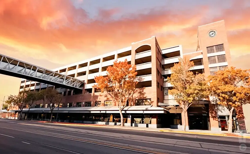 A multi-story brick building with a skybridge and autumn trees against a colorful sunset sky.