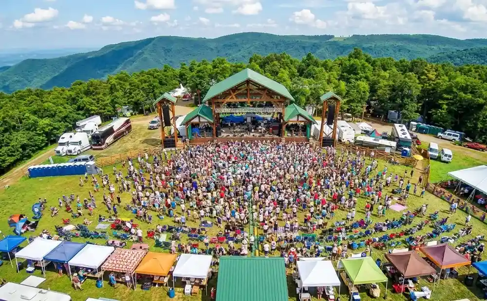 Aerial view of an outdoor music festival with a large crowd gathered in front of a stage, surrounded by tents and campers.