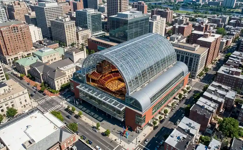 Aerial view of a large glass-domed concert hall with an organ and seating visible inside, surrounded by city buildings.