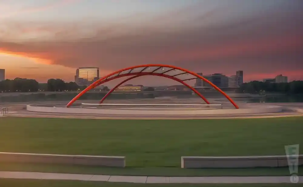 A sunset over a cityscape with a large orange arch sculpture in the foreground.