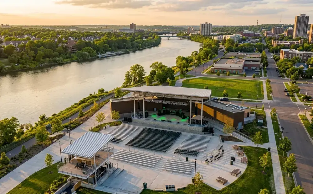 Aerial view of an outdoor amphitheater by a river, with a cityscape in the background at sunset.