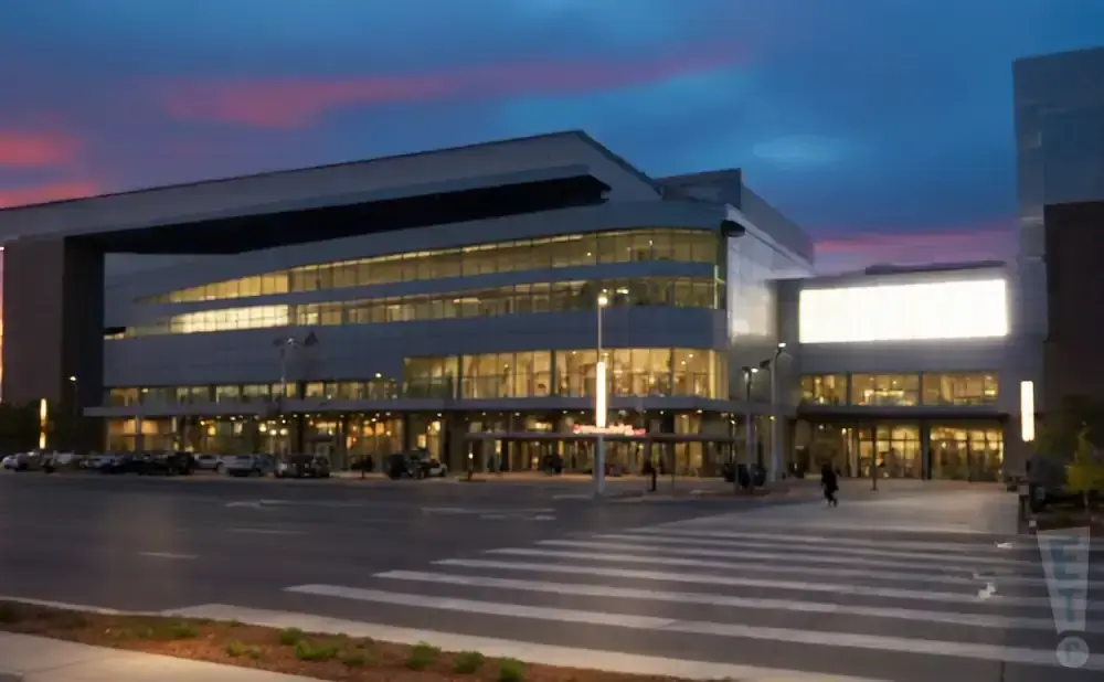 A modern convention center at dusk with a bright marquee and cars parked outside.
