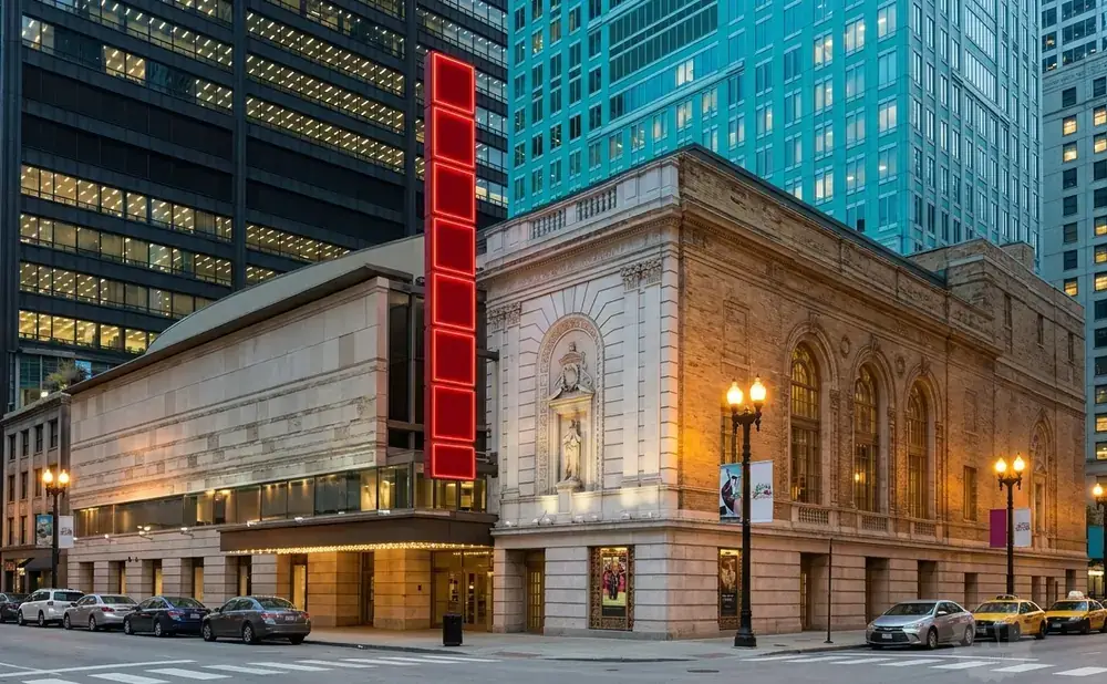 Theater with modern and historic architecture, neon sign, and taxis on a city street at dusk.
