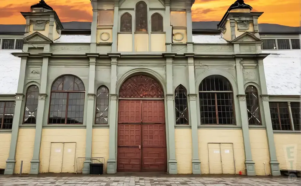 A grand, old building with a large red double door and arched windows under a dramatic orange sky.